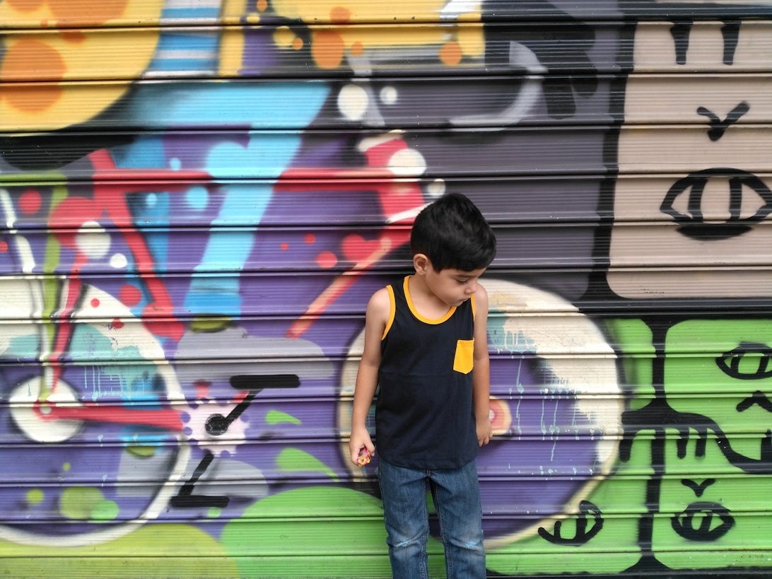 Een jong kind in een zwarte tanktop met gele details en een spijkerbroek poseert tegen een levendige muur vol graffiti, wat de stedelijke streetwear-esthetiek perfect weergeeft.